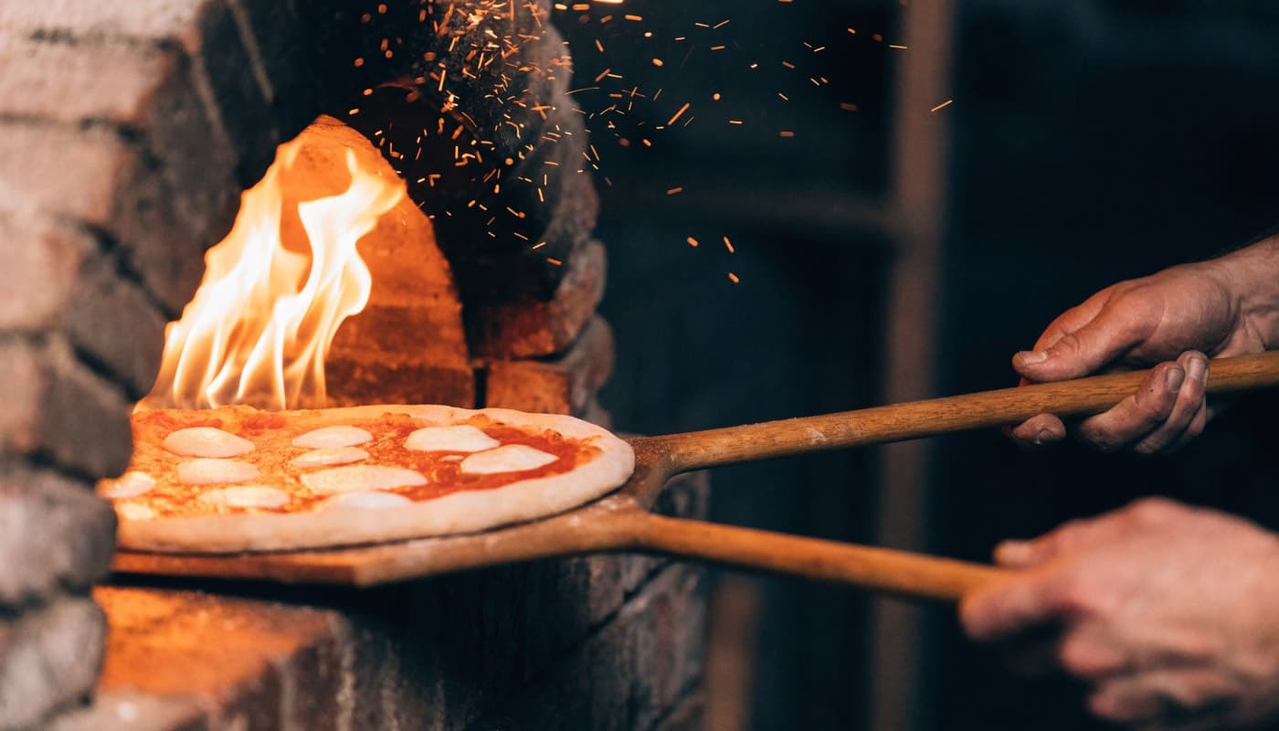 Pizza wordt met een houten schep in de vlammende mond van een steenoven geschoven